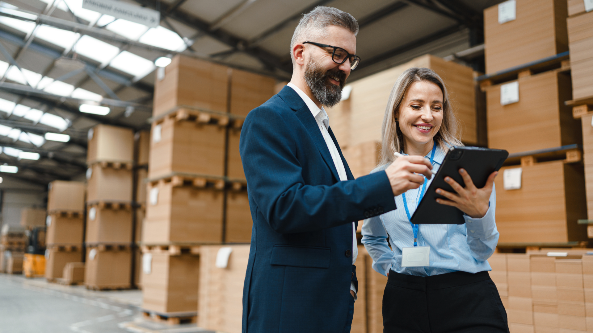A manager and employee review a tablet in a warehouse filled with stacks of cardboard boxes.