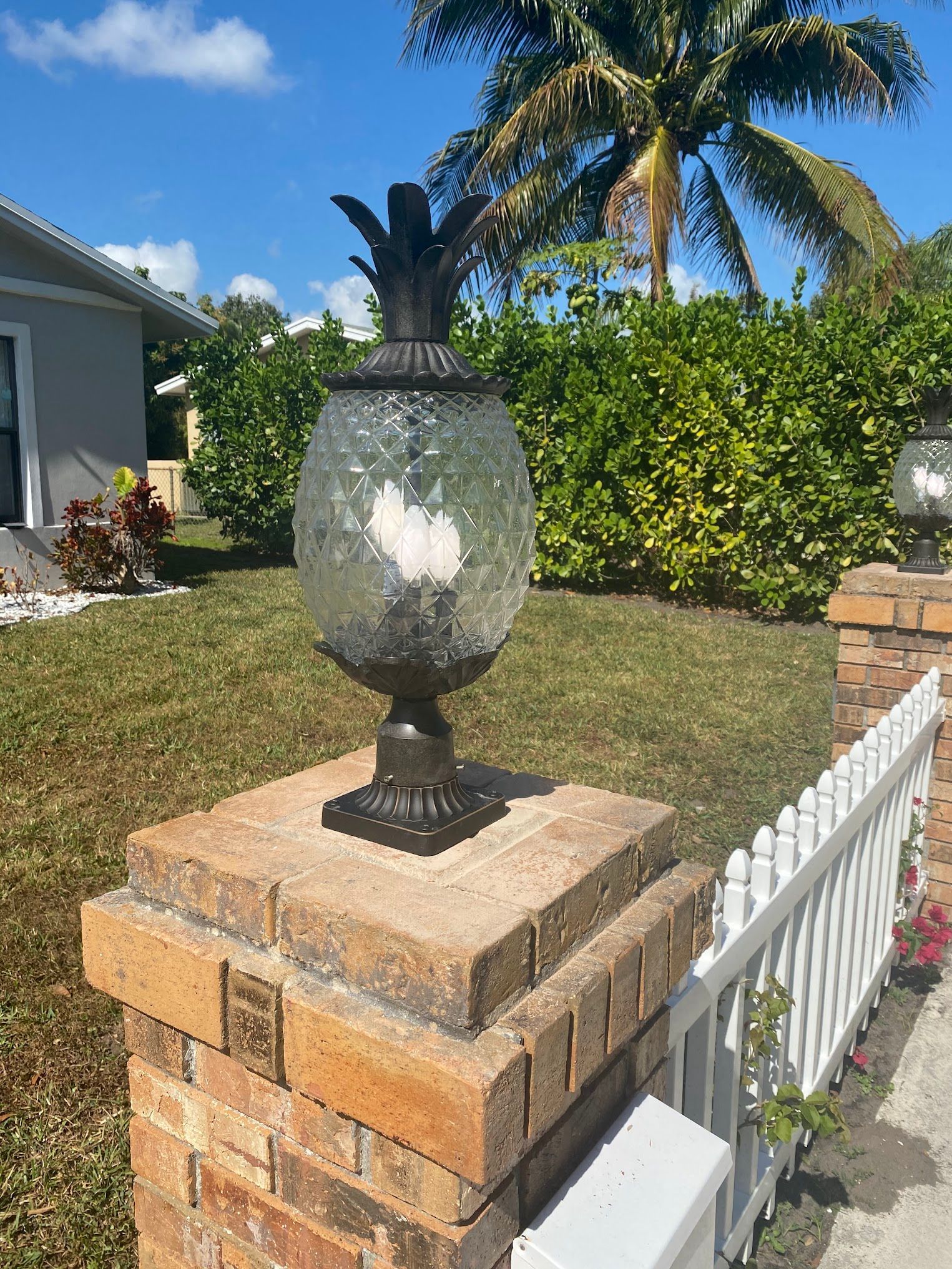 A lamp post in the shape of a pineapple is sitting on top of a brick post next to a white picket fence.