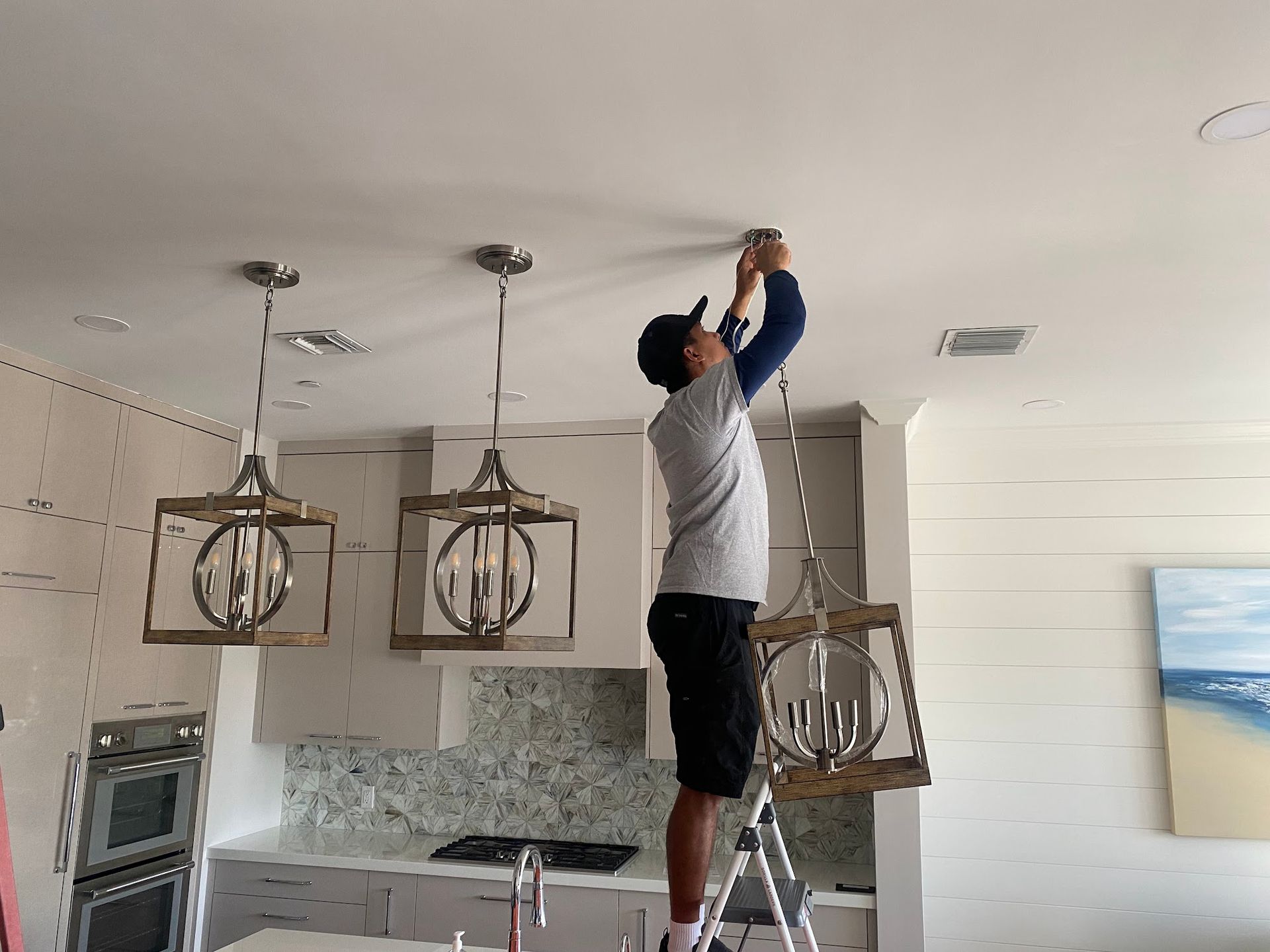 A man is standing on a ladder fixing a light fixture in a kitchen.