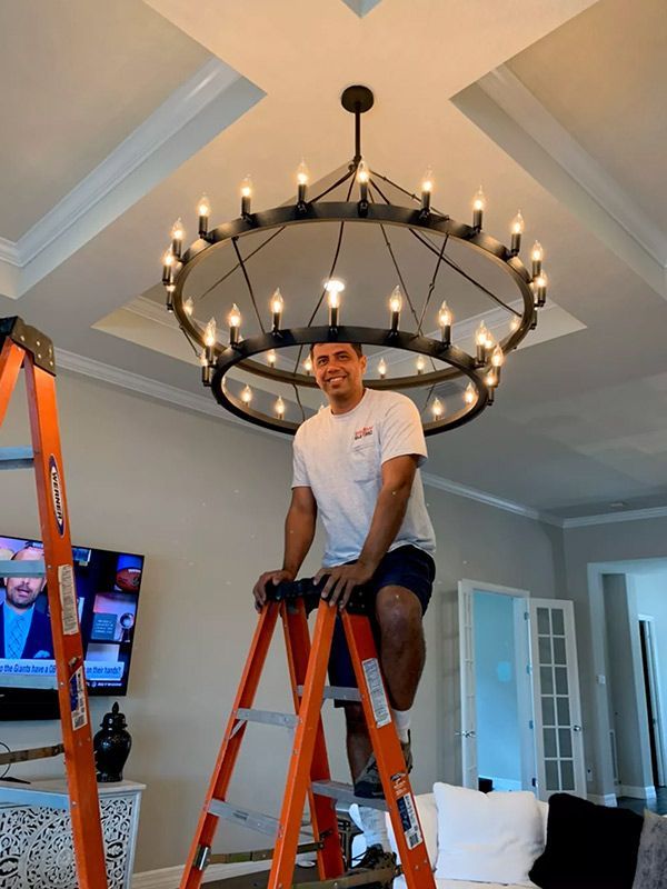 A man is standing on a ladder in front of a chandelier in a living room.