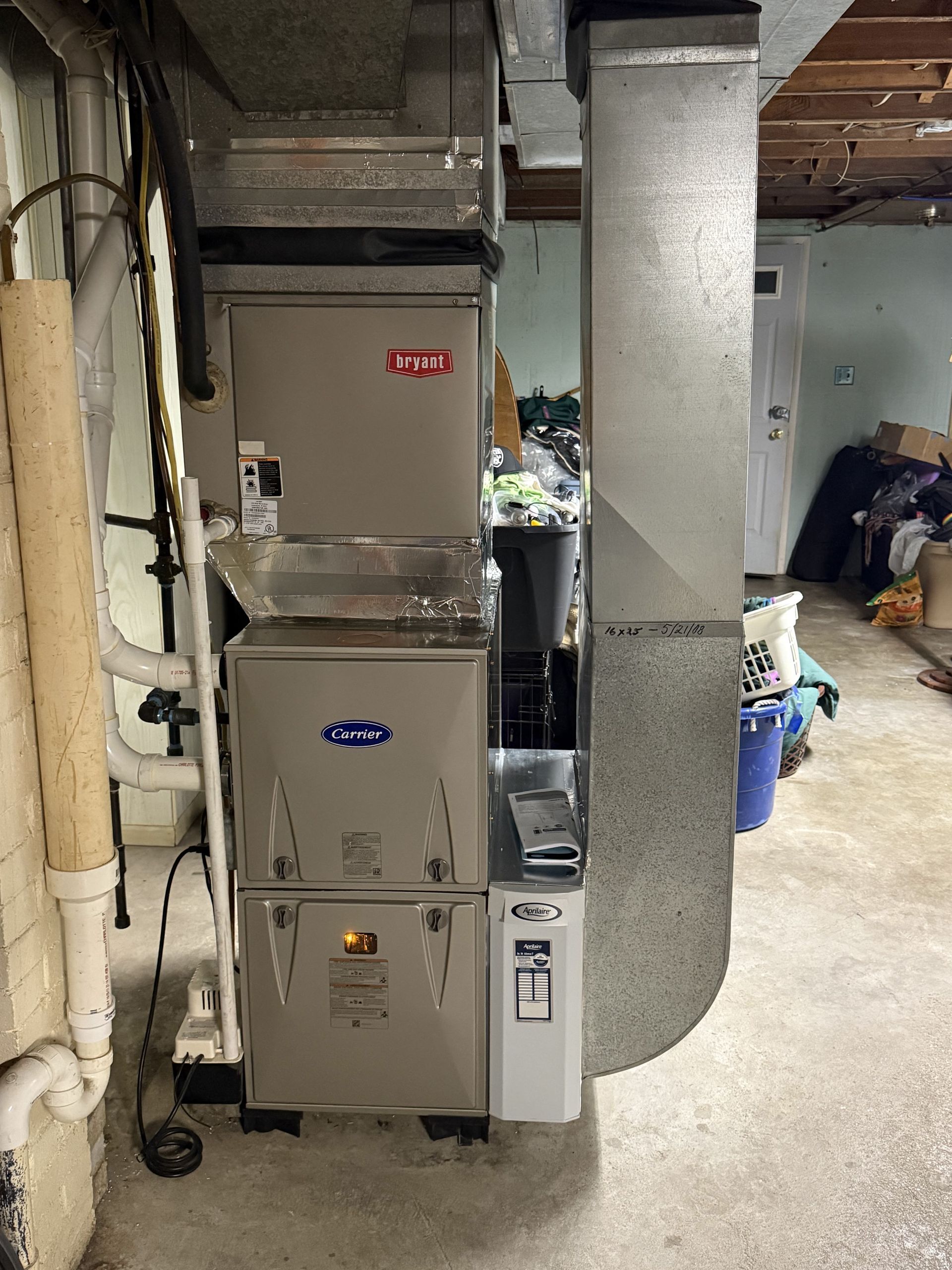 A metallic HVAC furnace unit stands in a basement next to a vertical silver air duct, surrounded by storage items.