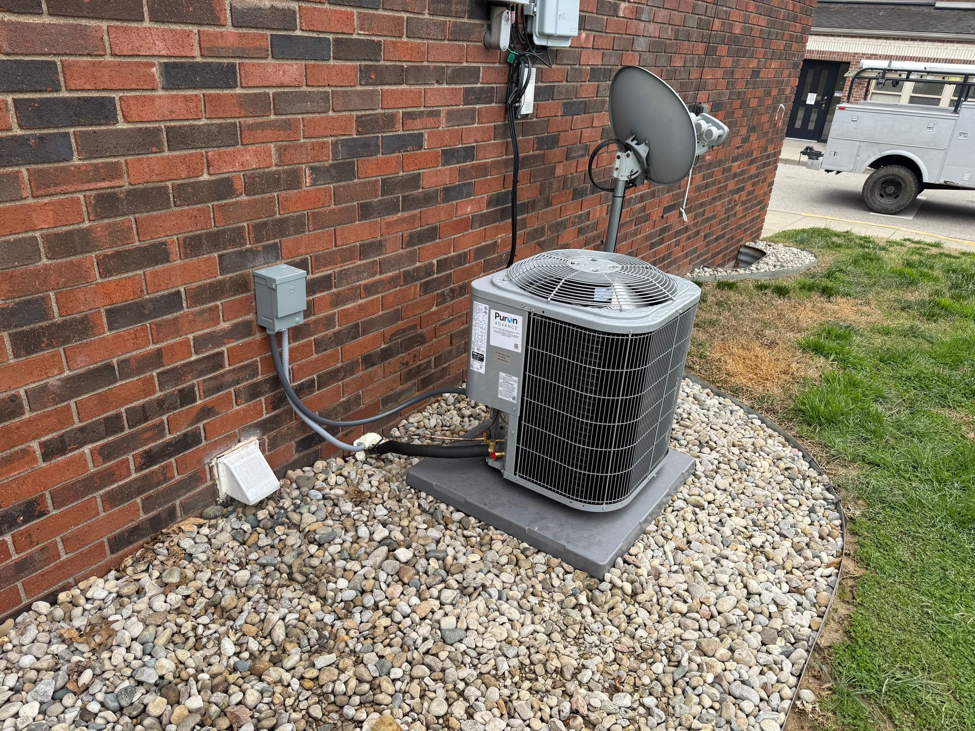 An outdoor air conditioning unit sits on a concrete pad against a brick wall next to a satellite dish and electrical box.