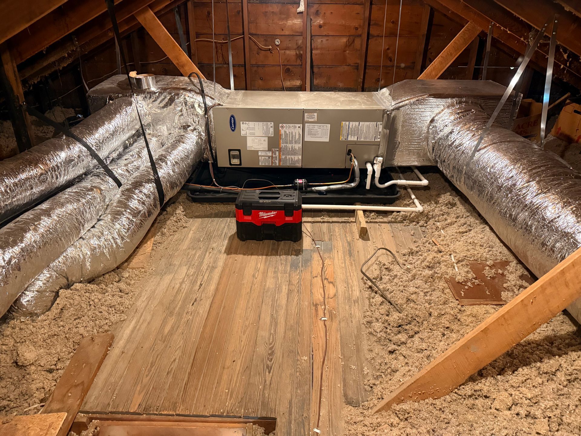 An attic-mounted HVAC unit with insulated ductwork, resting on a black drain pan above loose-fill insulation.