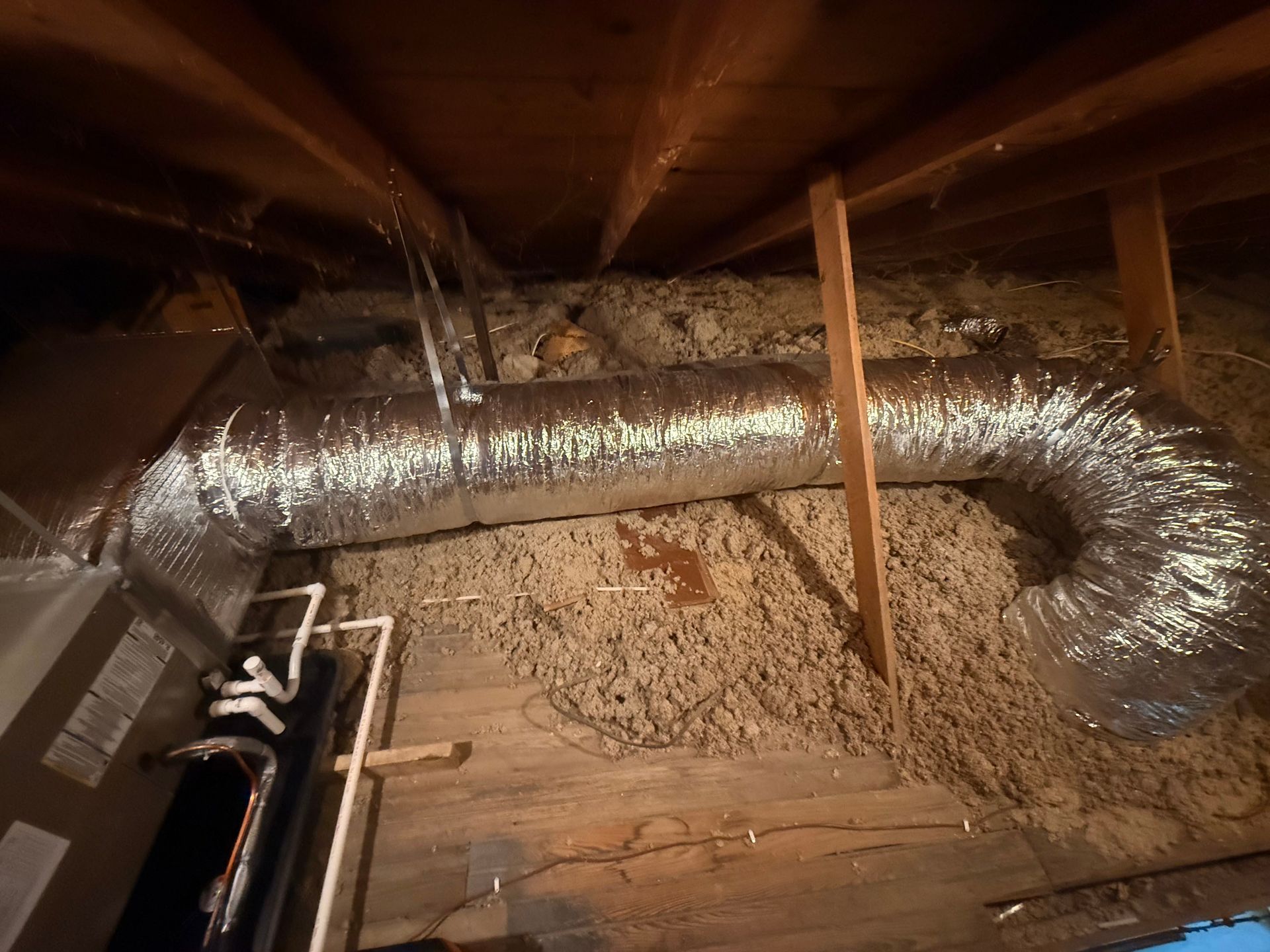 A view inside an attic, showing an HVAC unit connected to a long, insulated flexible duct laying over loose insulation.