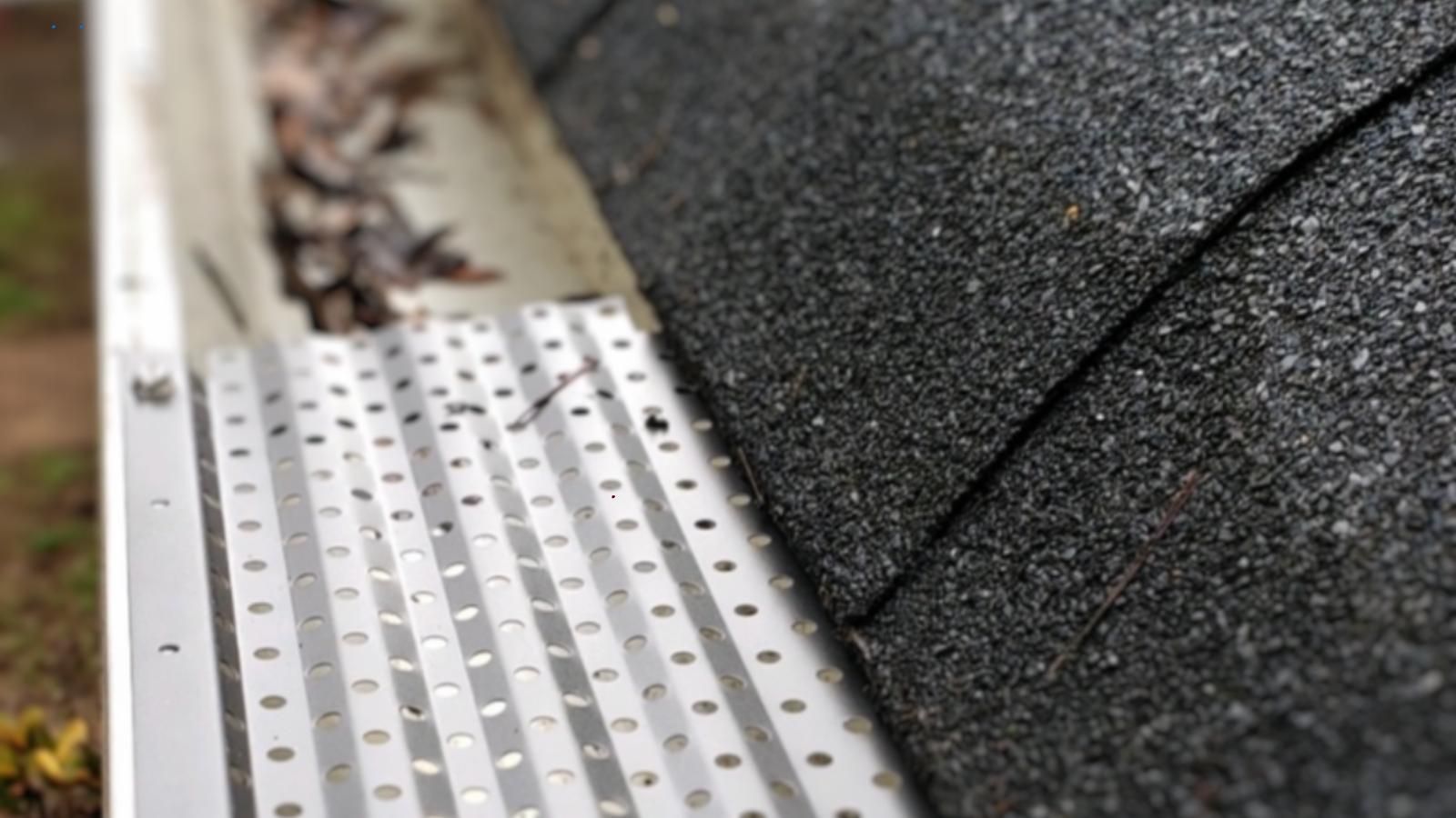 A close up of a gutter on a roof with leaves coming out of it. Part of the gutter has a guard installed.