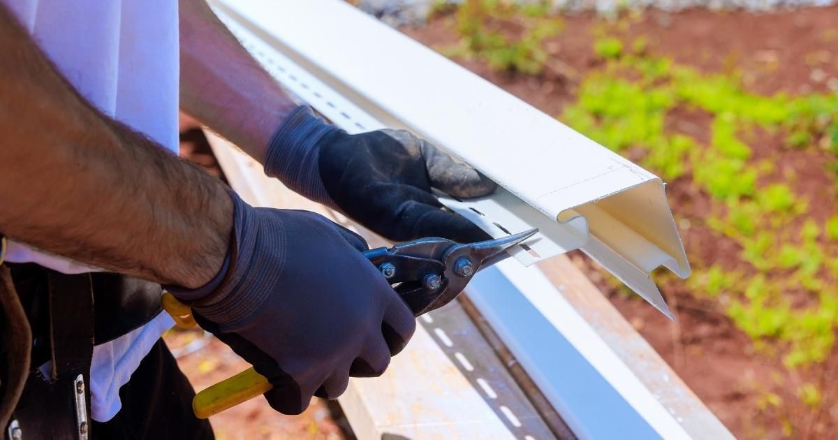 A worker wearing black gloves uses tin snips to trim a white vinyl siding piece outdoors.