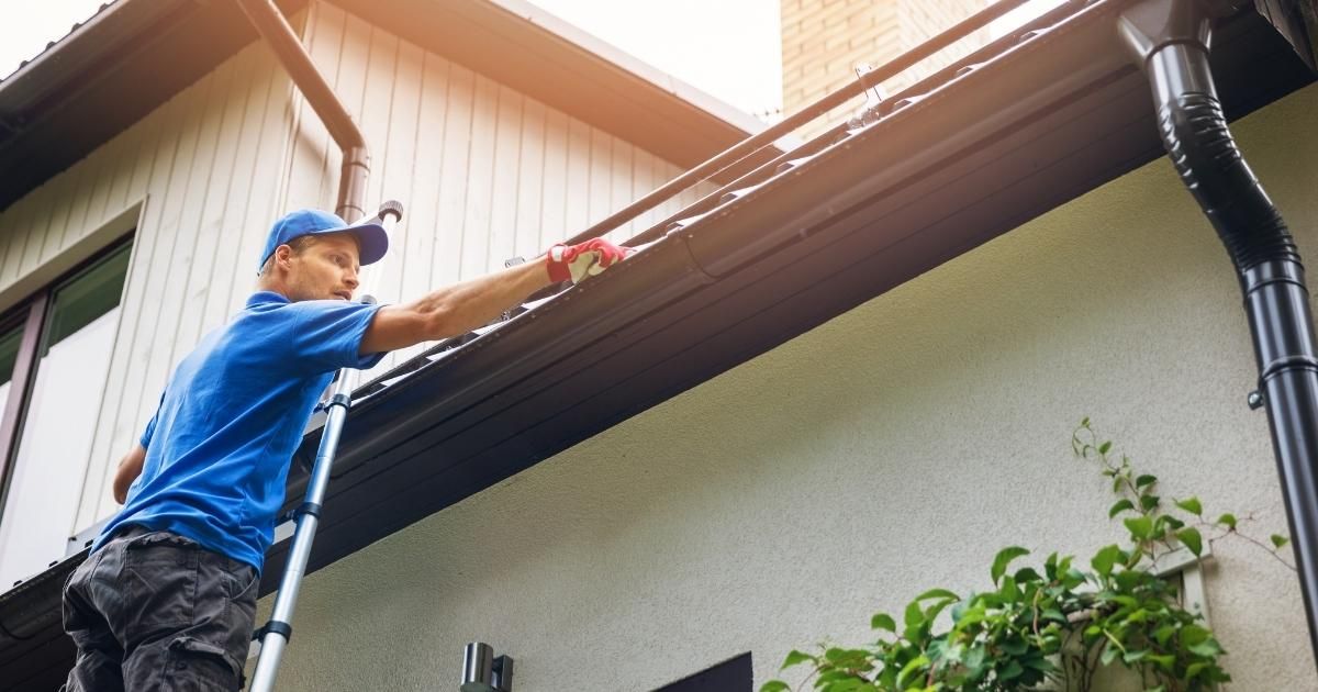 A worker in a blue shirt and cap stands on a ladder cleaning debris from a brown house gutter.