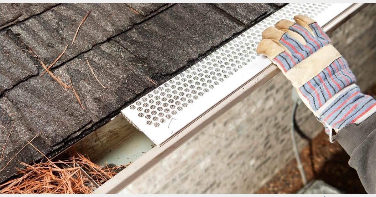Gloved hand cleaning gutter with mesh guard, removing debris from roof.