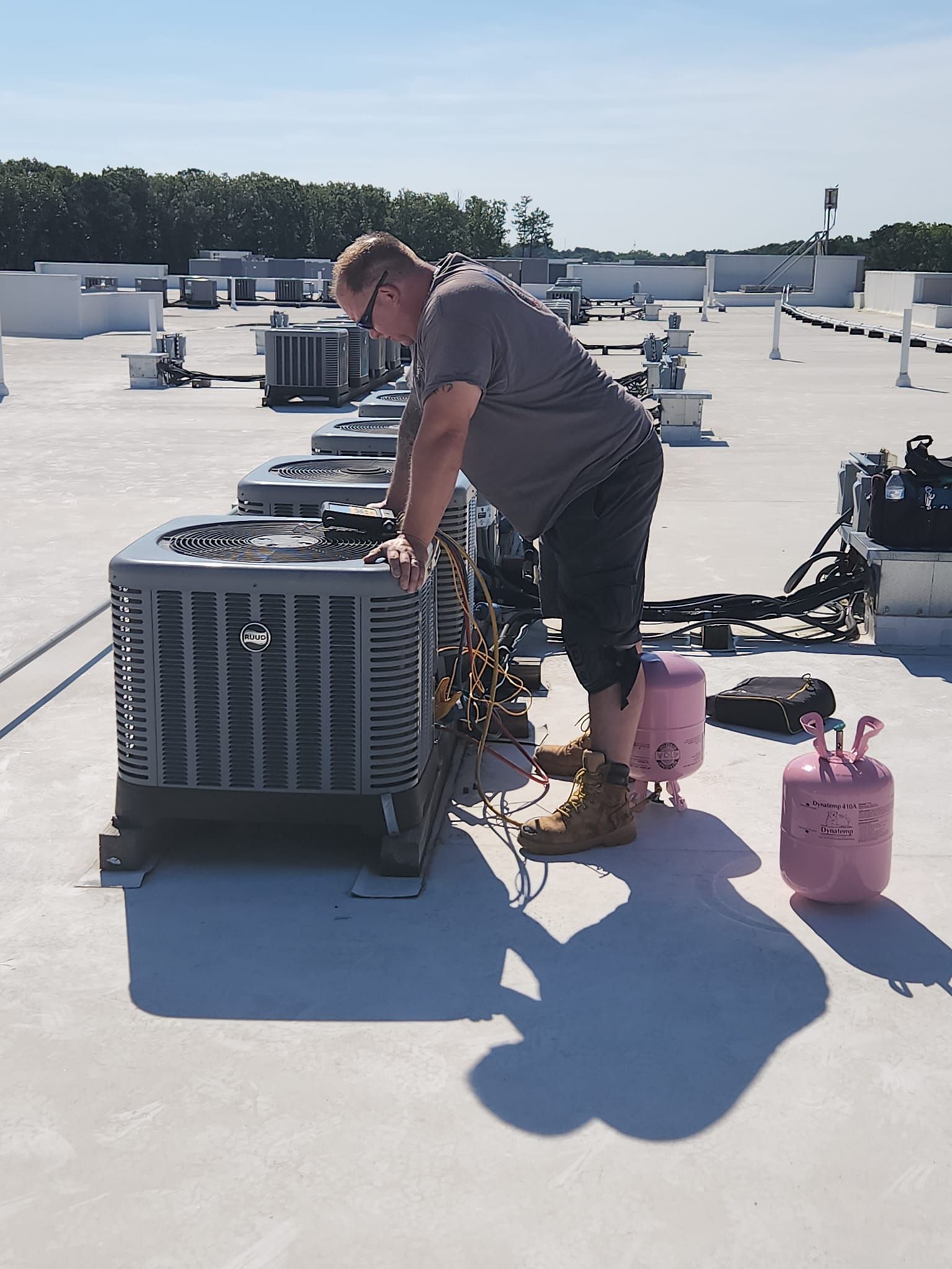 Man in gray shirt works on HVAC unit on a rooftop under a sunny sky, next to pink tanks.