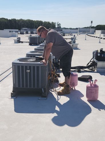 Man in gray shirt works on HVAC unit on a rooftop under a sunny sky, next to pink tanks.