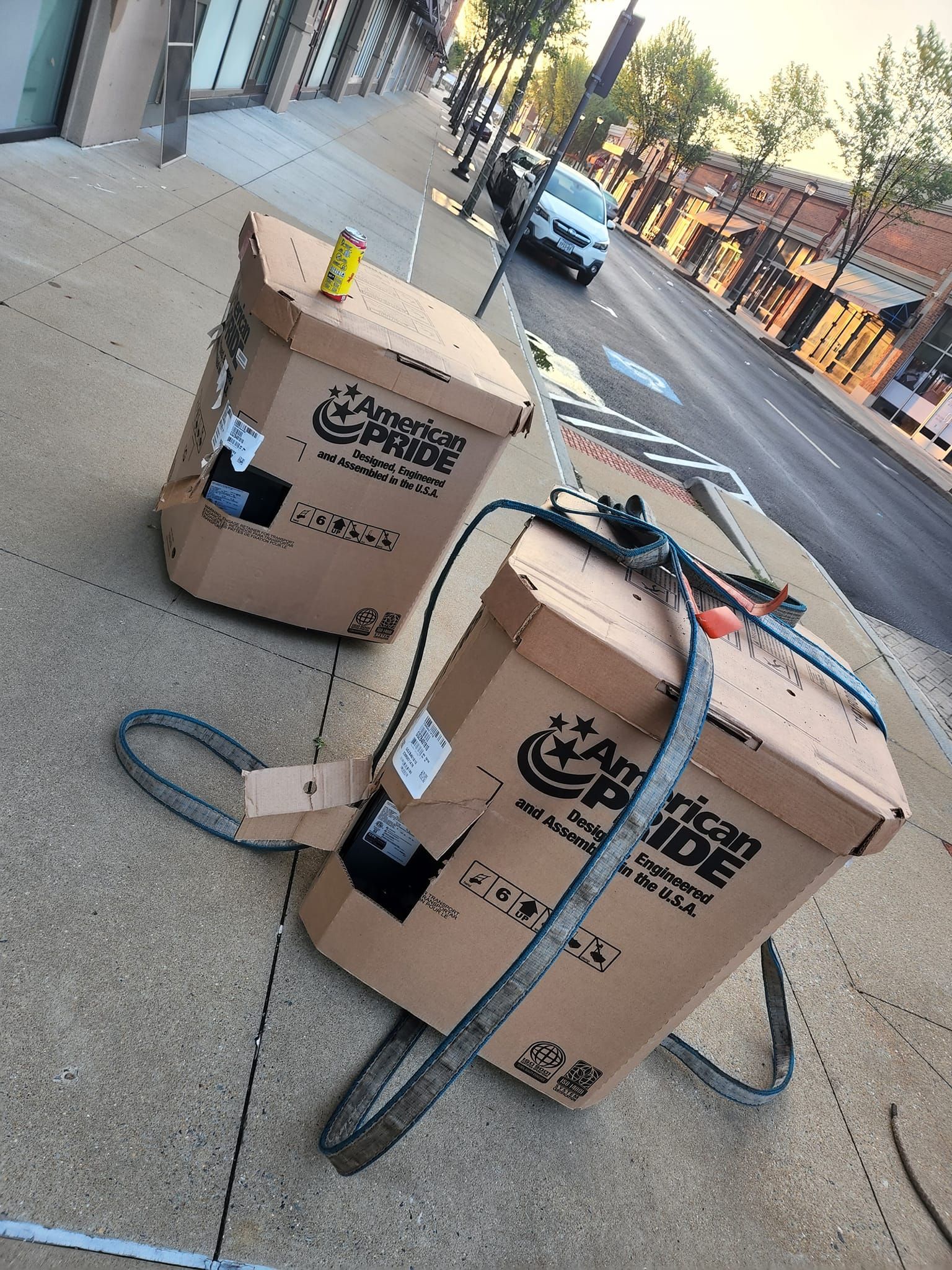 Two cardboard boxes with straps on a sidewalk. One has a yellow object on top.