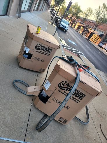 Two cardboard boxes with straps on a sidewalk. One has a yellow object on top.