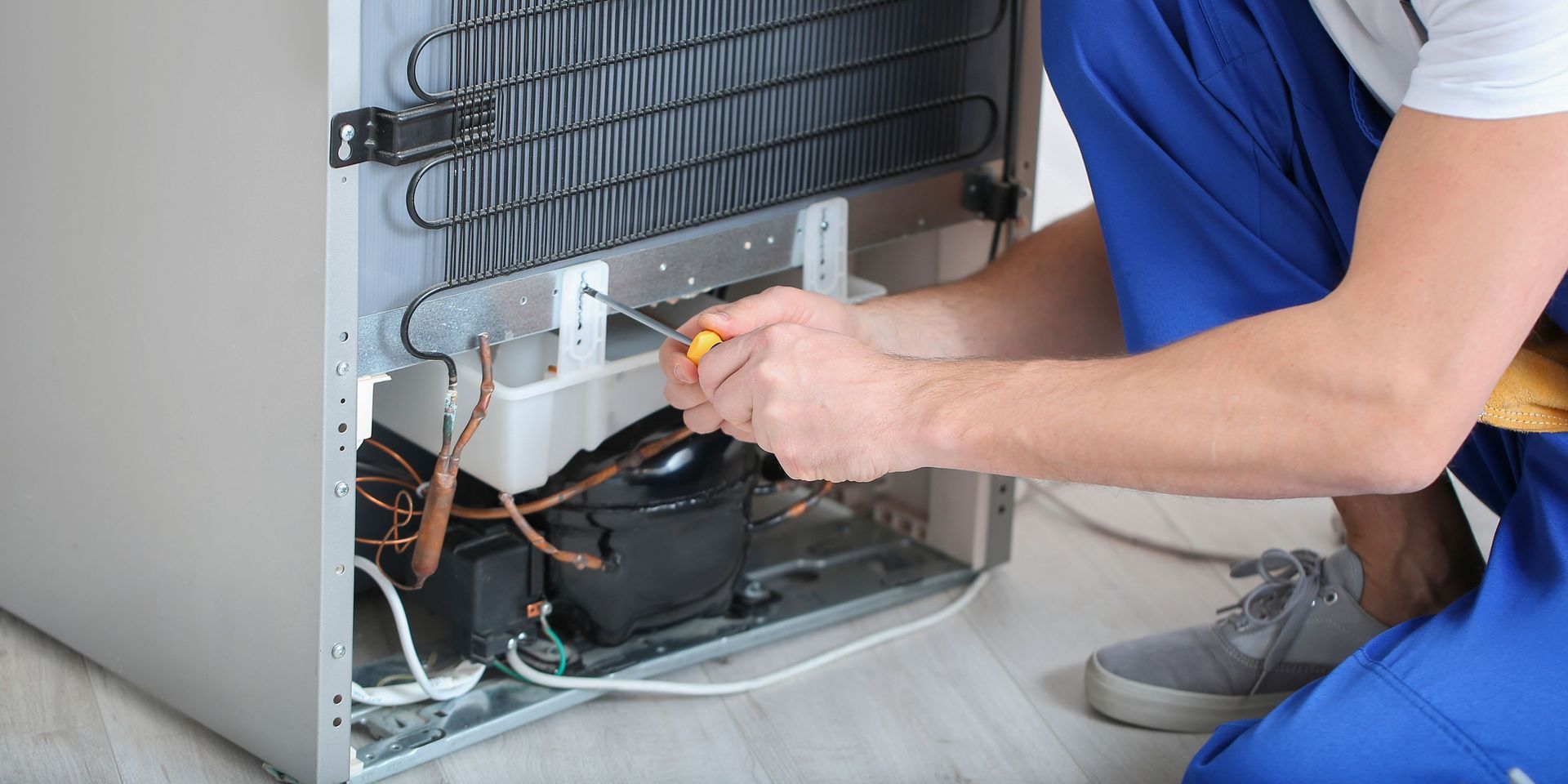A person in blue overalls repairs a refrigerator, using a screwdriver.