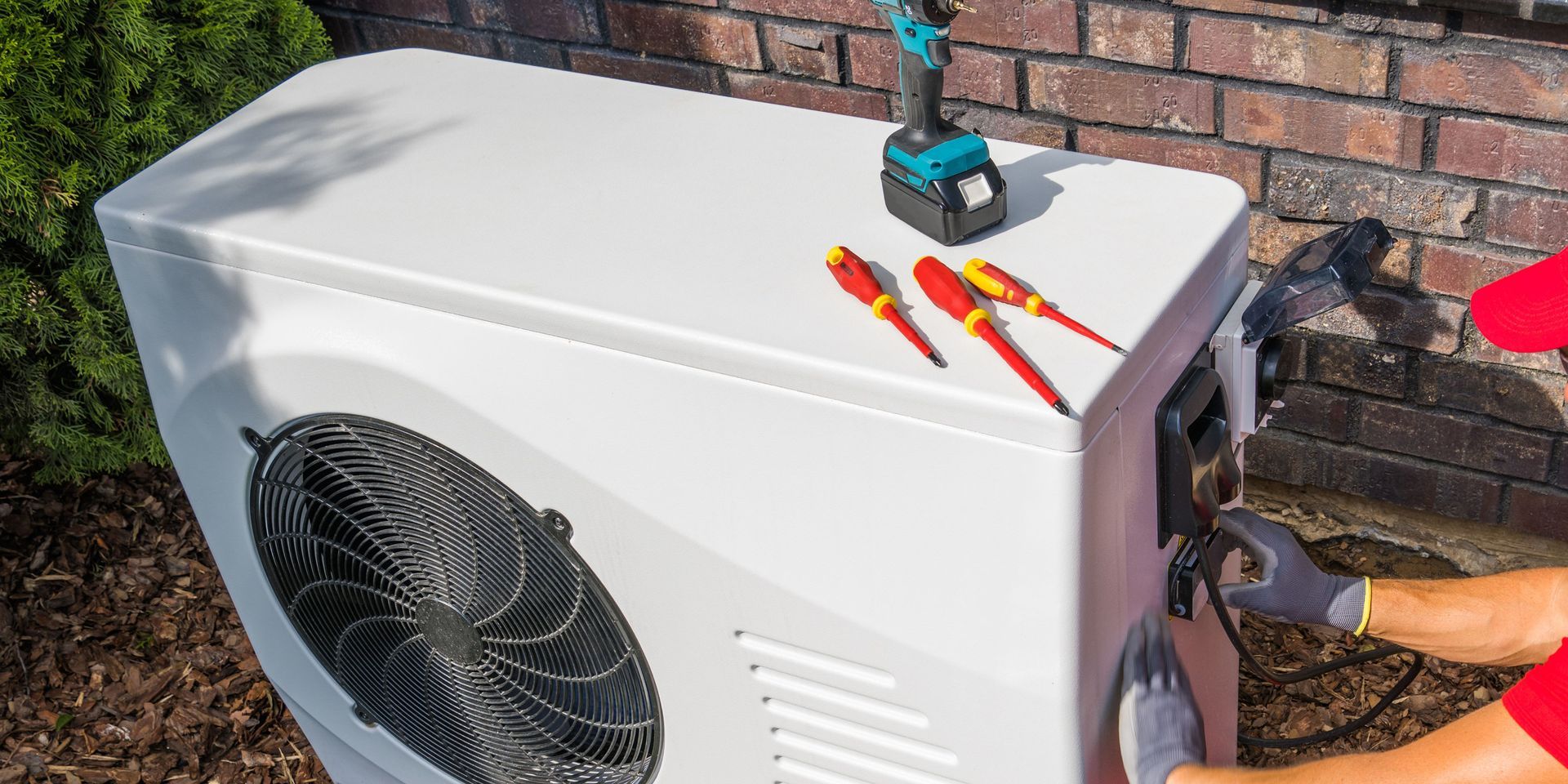 Person in gloves repairs a white air conditioner with tools on top, near brick wall and green bushes.