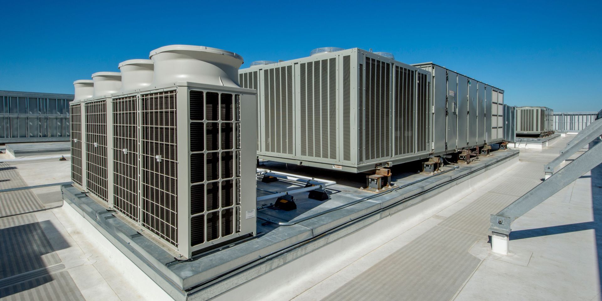 Rooftop HVAC units on a bright, sunny day. Metal structures with vents, surrounded by a white surface and blue sky.