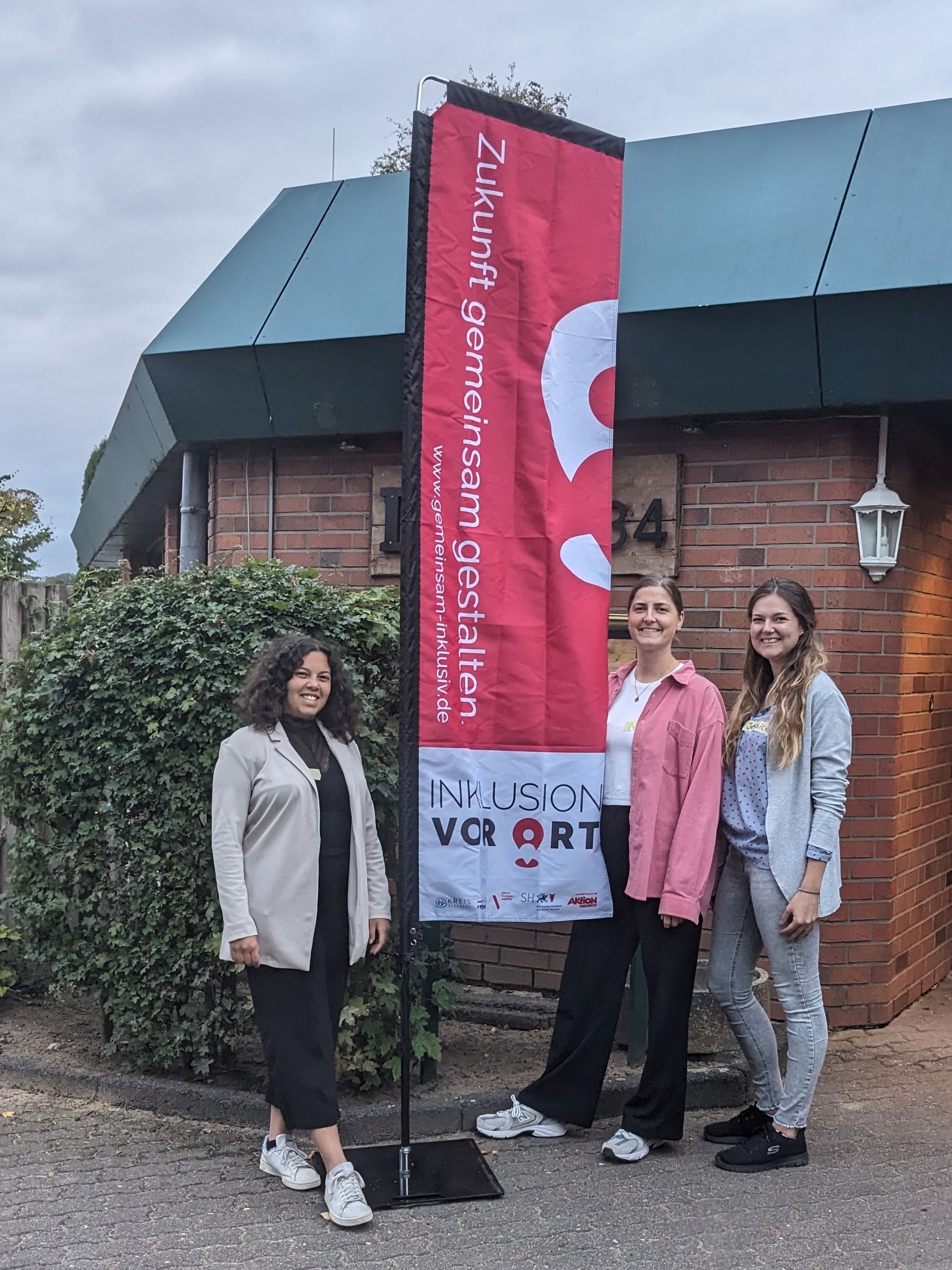 Auf dem Bild sind drei junge Frauen draußen vor einem Haus zu sehen, die den Workshop durchführen. Alle drei lächeln und stehen neben einer großen Fahne mit dem Projektlogo.