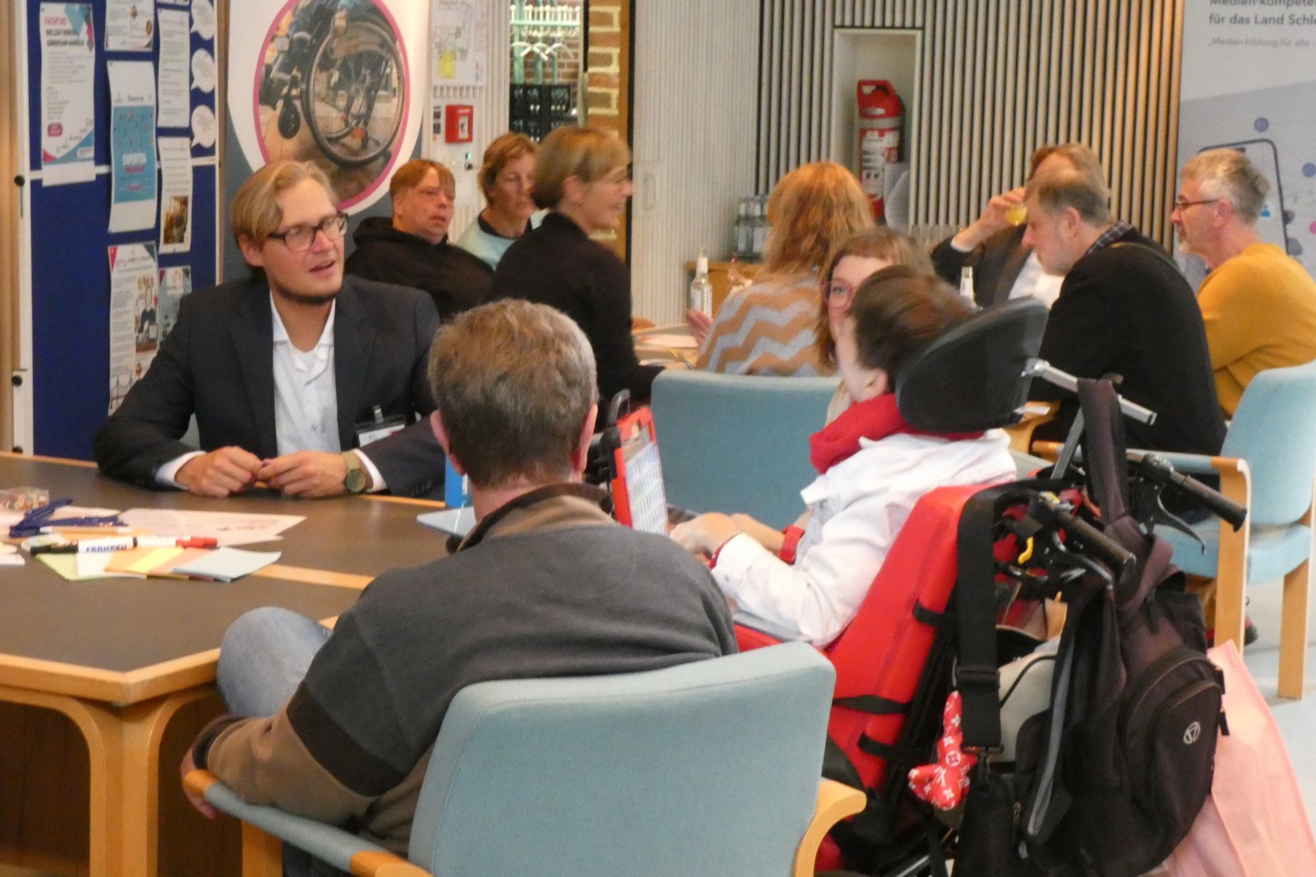 Eine Gruppe von menschen mit und ohne Behinderungen sitzen an einem Tisch und reden.
