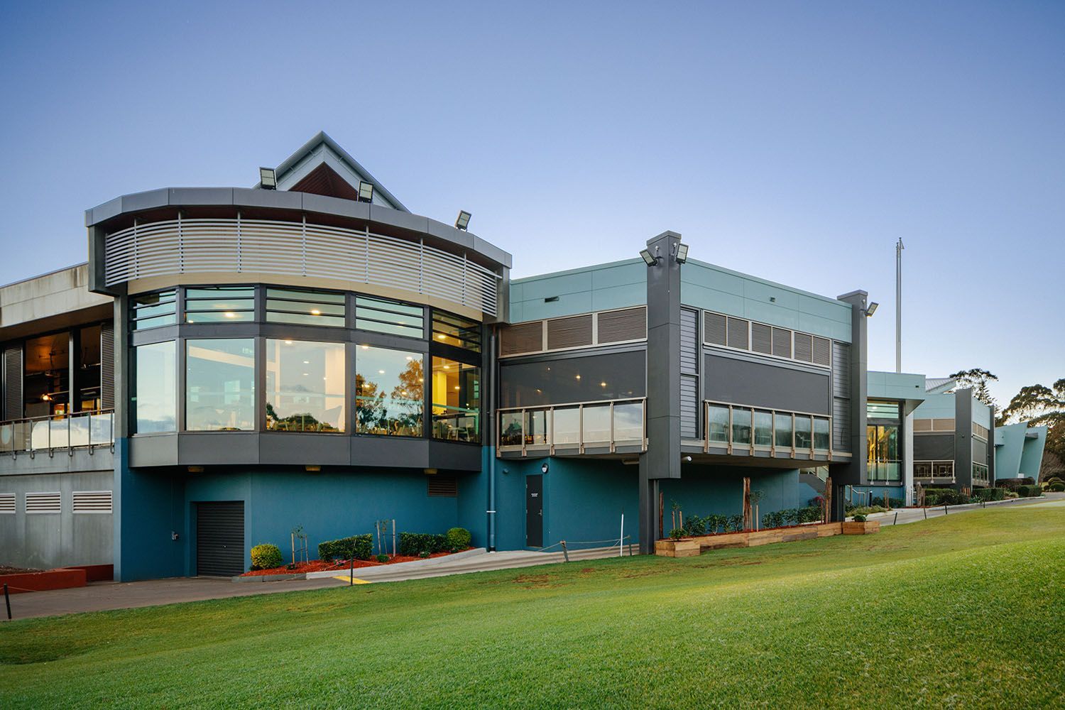 Modern building with curved glass windows and blue and gray exterior, viewed from a grassy lawn.