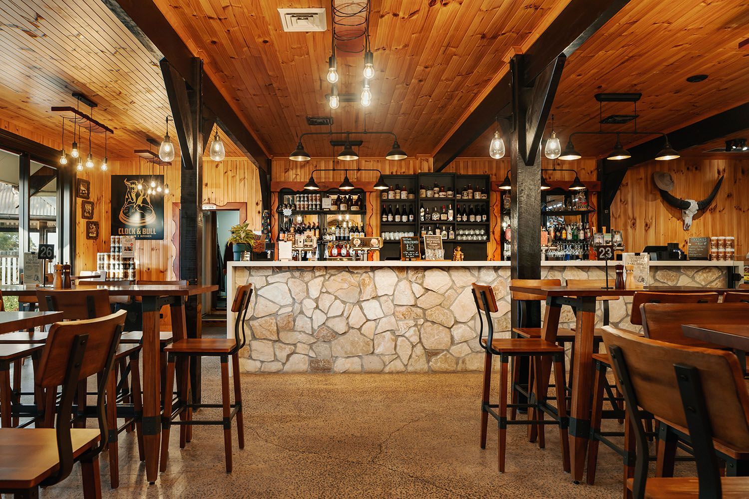 Interior of a rustic bar with wooden beams, stone bar front, tables, and stools.