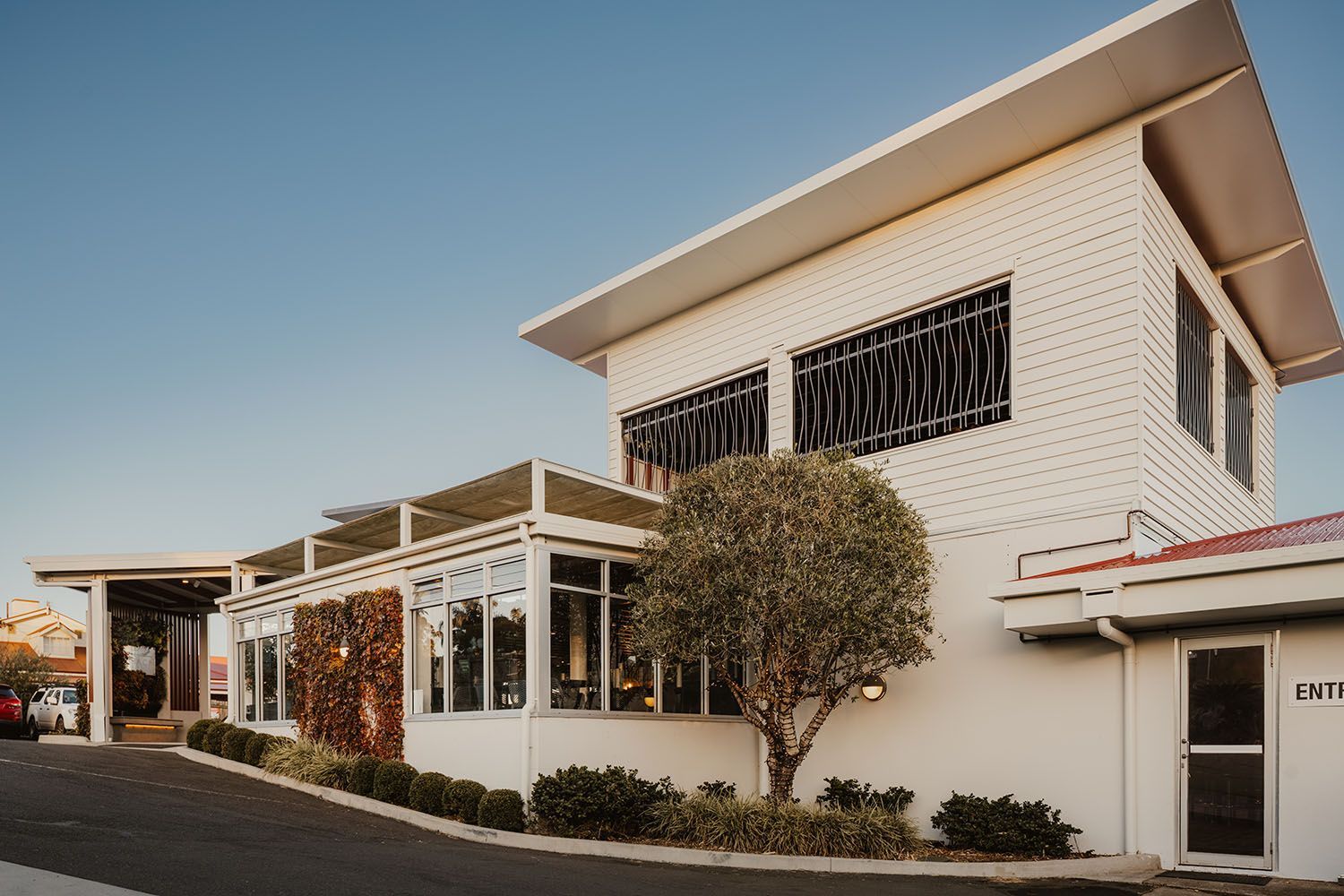 White building with a flat roof, large windows, and a small tree in front.