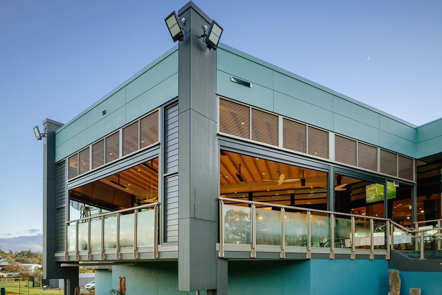 Modern, two-story building with blue exterior and wooden accents. Glass railing, spotlights, and open-air seating.