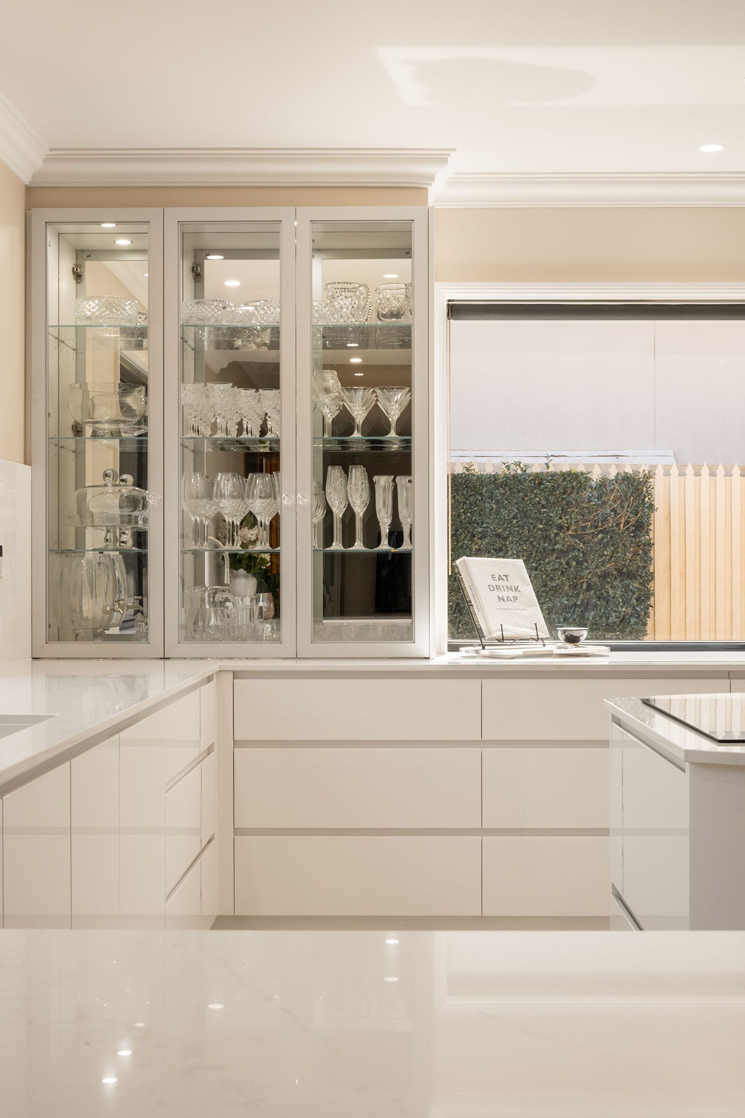 White kitchen with glass-front cabinets displaying glassware, countertop, and window with outdoor greenery.