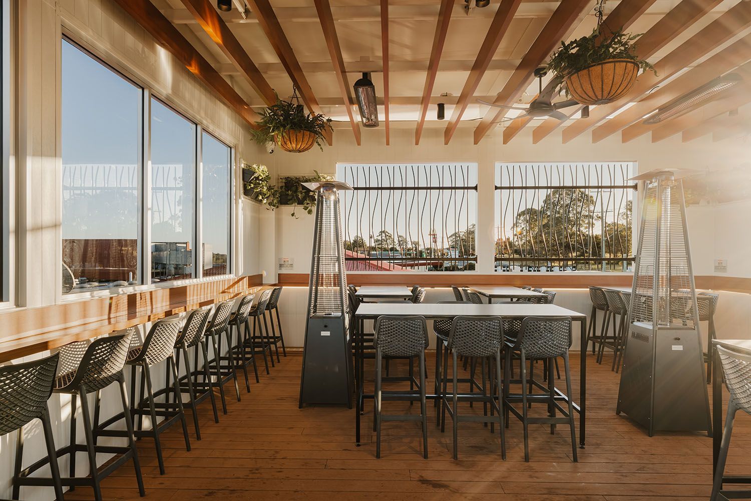Rooftop bar with wood floor, tall windows, and a lattice wall. Tables, chairs, and hanging plants are visible.