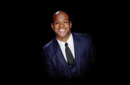 Man in a navy suit with a black tie smiles against a black background.