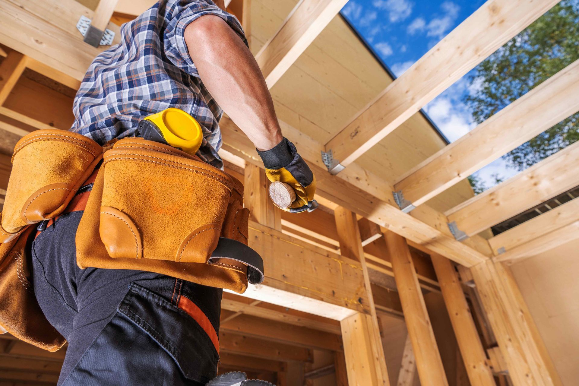 Construction worker installing ceiling joists in a wooden house frame, wearing a tool belt and gloves