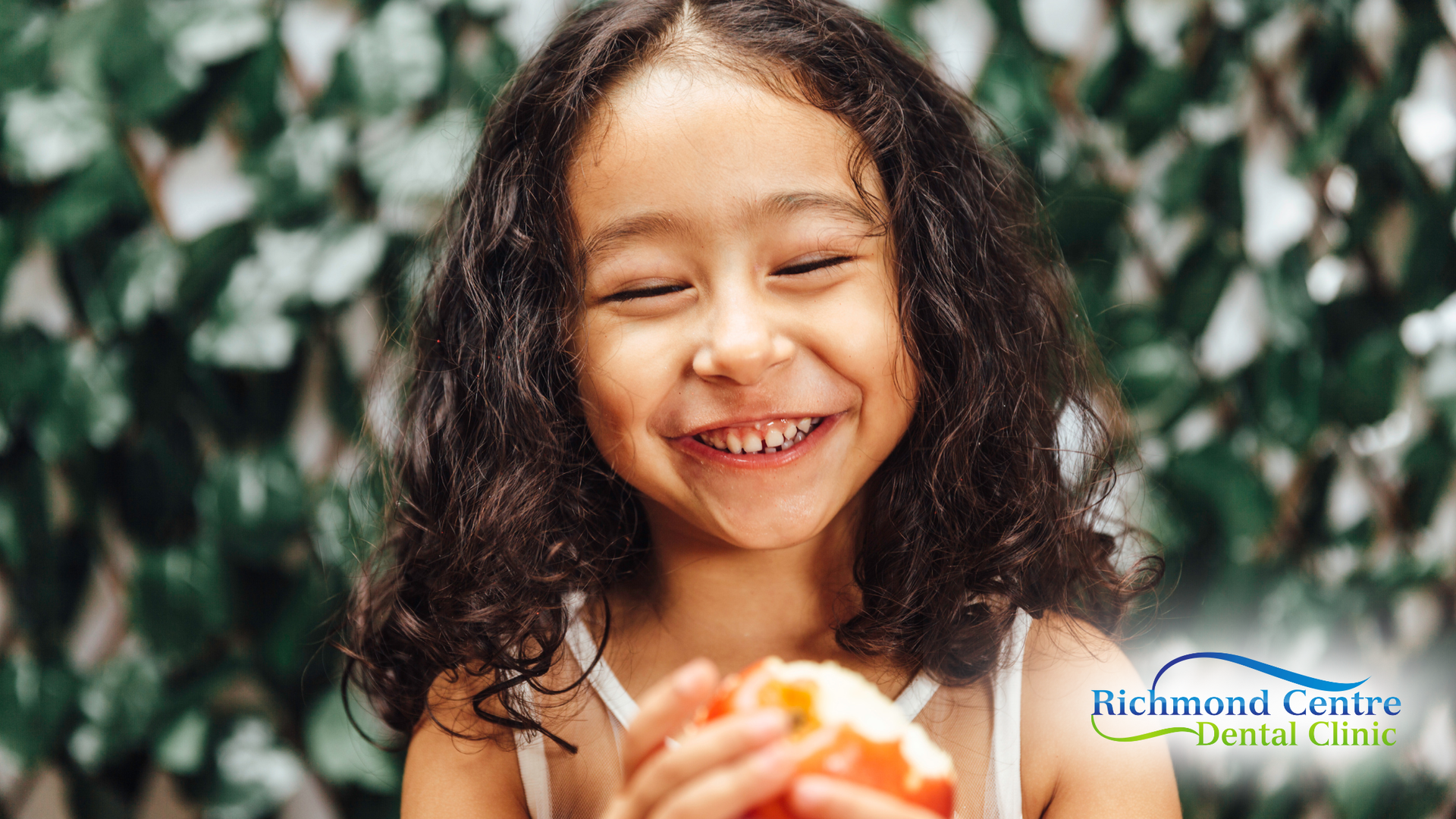 Smiling child holding a piece of fruit. Green foliage in background.