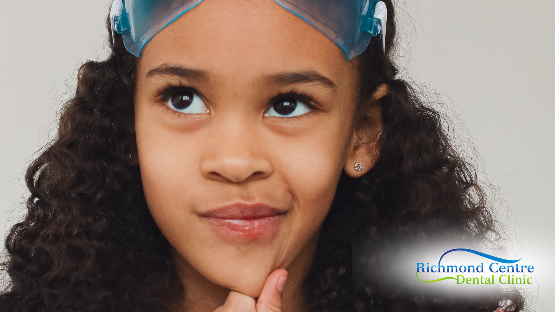 Girl with swim goggles on forehead looking up thoughtfully, hand on chin.