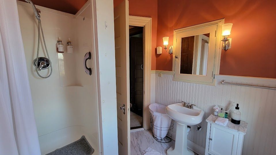 Bathroom with white shower and sink, orange wall, mirror with sconces, and open door.