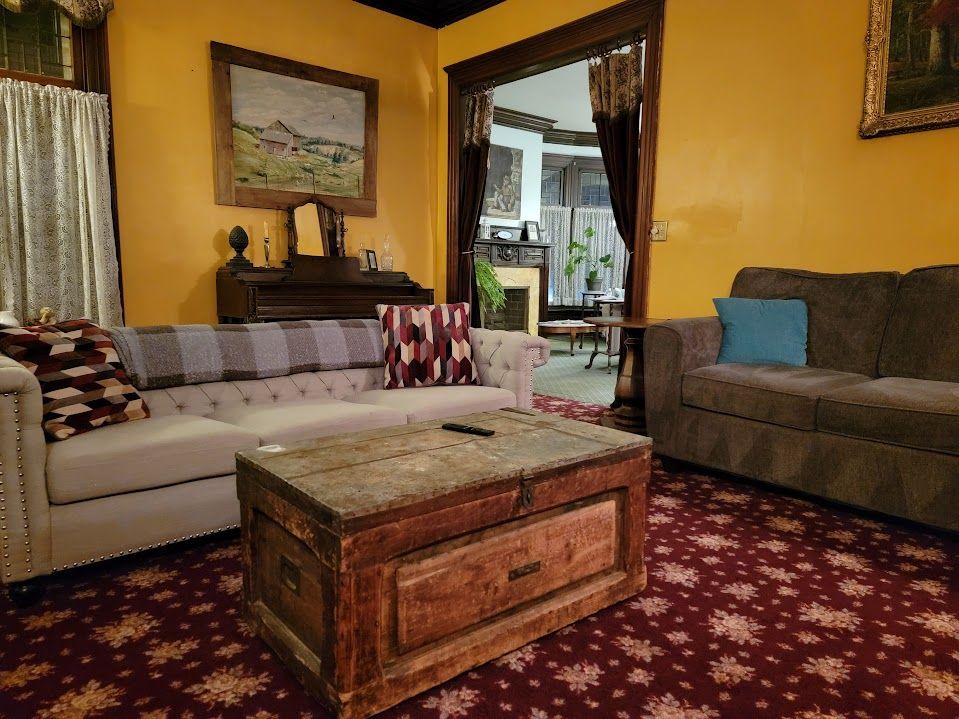 Living room with a tufted sofa, wooden chest, and a doorway to another room. Yellow walls and patterned rug.