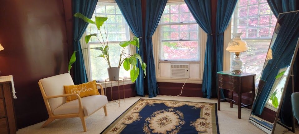 Cozy room with teal curtains, a chair with a yellow pillow, and a rug. The room has large windows and a floor mirror.