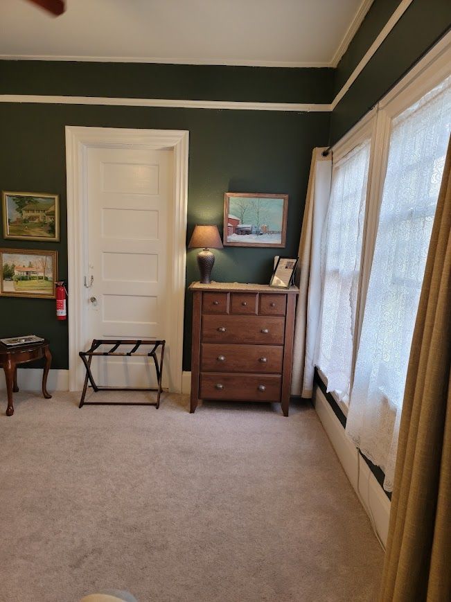 Room with dark green walls, wood dresser, artwork, door, carpet, and window with curtains.