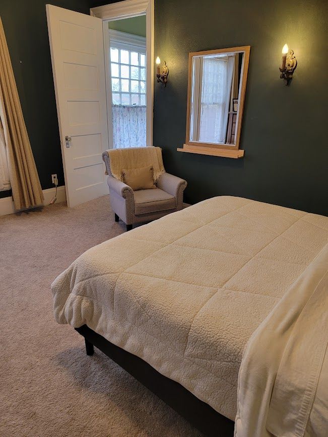 Bedroom with a bed, armchair, mirror, and sconces on a dark green wall; doorway leads to an outdoor space.