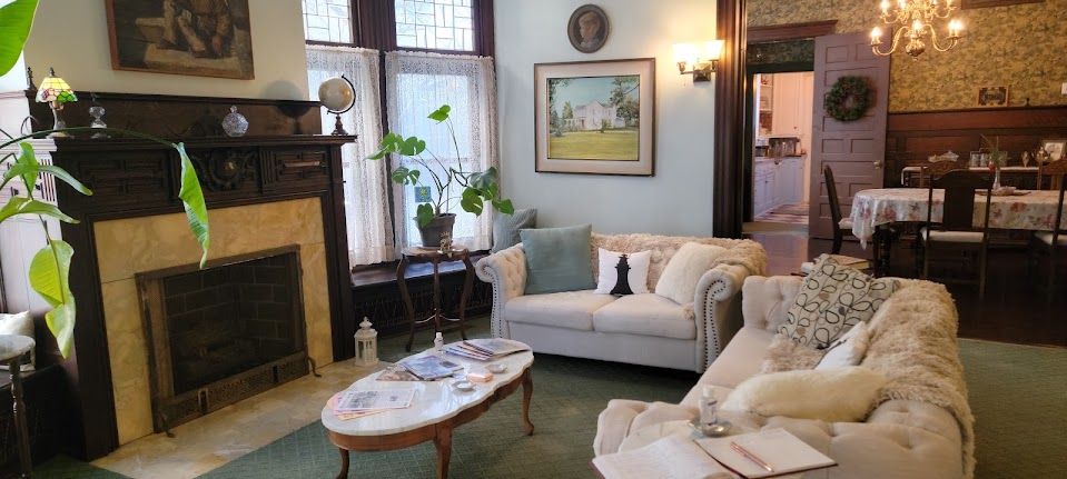 Living room with fireplace, white couches, and a dining area in the background. Green carpet, floral wallpaper.