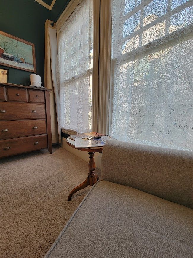 A bedroom corner with a dresser, small table with books, window with light filtering through sheer curtains.