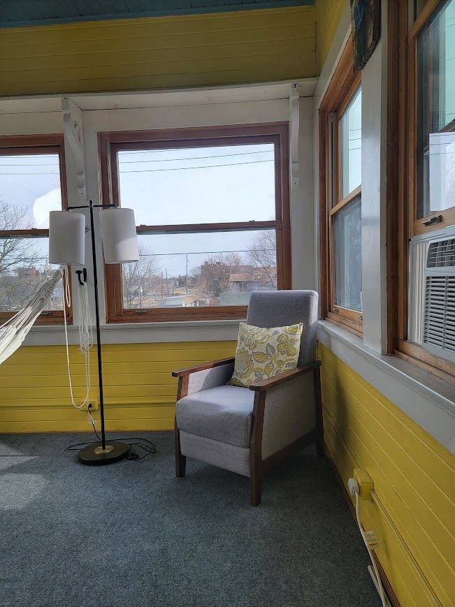 Sunroom with gray armchair, floor lamp, yellow walls, and windows overlooking a leafless landscape.