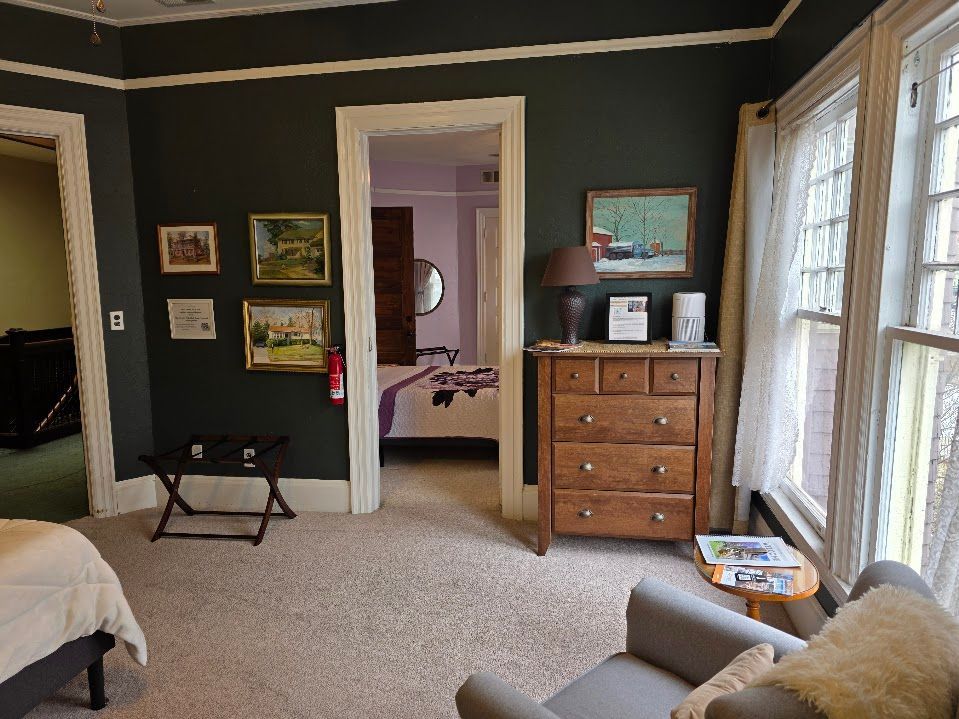 Bedroom with dark green walls, dresser, art, doorway to another room, window with sheer curtains, and a reading chair.