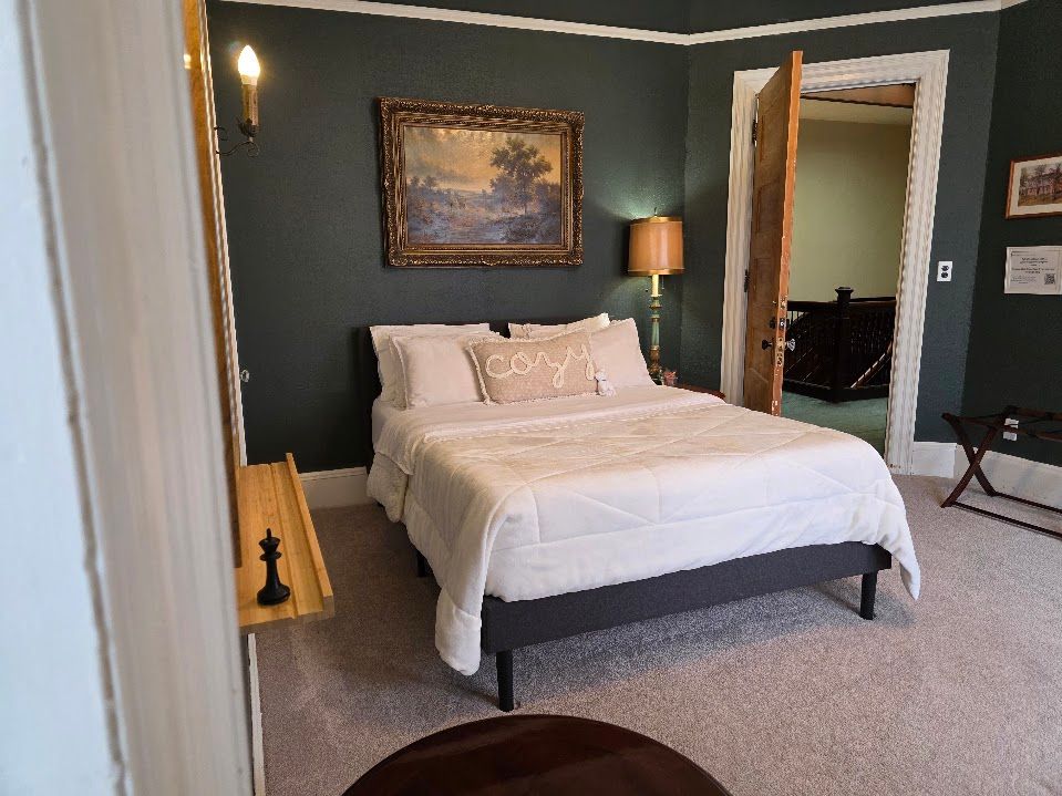 Bedroom with a made bed, dark walls, a painting, and an open doorway to another room.