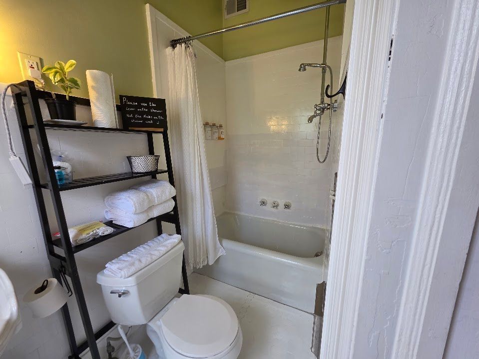 Small bathroom with a white tub and toilet, shower curtain, and a black shelving unit with items.