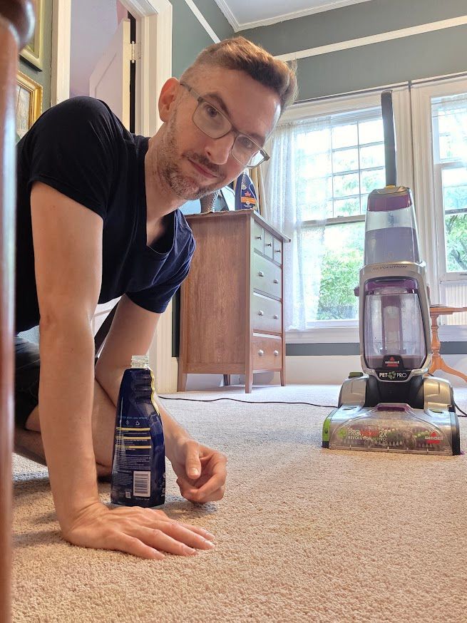 Man kneels on carpet with vacuum. He looks at the camera, holding a blue bottle. A dresser and window are in the background.