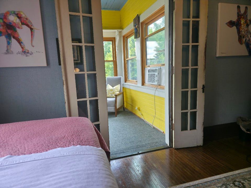 Bedroom with open doors leading to a sunroom; pink bedspread, gray walls, yellow sunroom walls, art on walls.