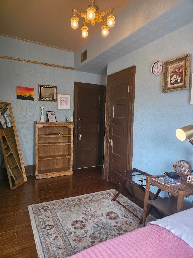 Cozy bedroom with light blue walls, wood floors, and vintage furniture. Includes a rug, bookshelf, and decorative art.