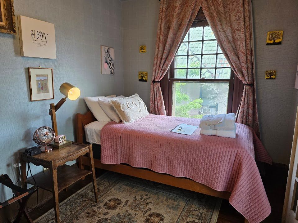 Bedroom with pink bedspread, wooden furniture, artwork, and window with floral curtains.