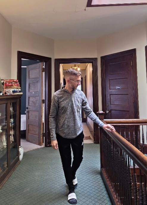 Man walking down a hallway, holding a railing.  Gray and black clothing, interior, and wood doors.
