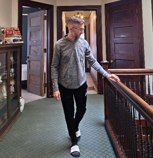 Man walking down a hallway with closed doors, wearing a gray hoodie and black pants.