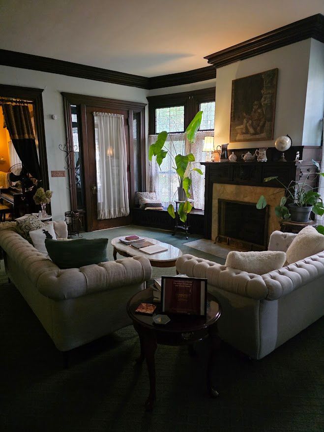 Cozy living room with tufted sofas, fireplace, and large windows. Dark wood trim and plants add warmth.