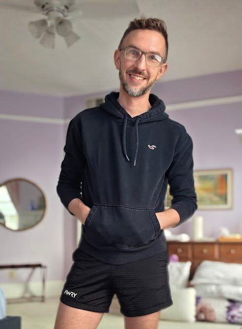 Man in glasses, blue hoodie, and black shorts, smiling, hands in pockets, standing in a room.